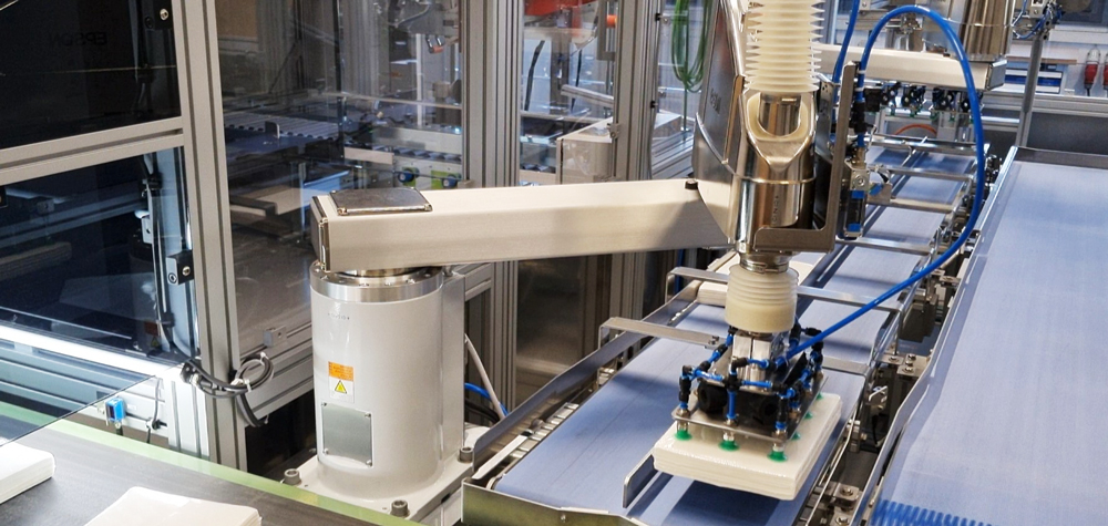 Automatisierte-verpackungsanlage-fuer-maximale-flexibilitaet-item-blog-bild-im-beitrag-2 Un robot con pinzas depositando un paquete de papel en una cinta transportadora
