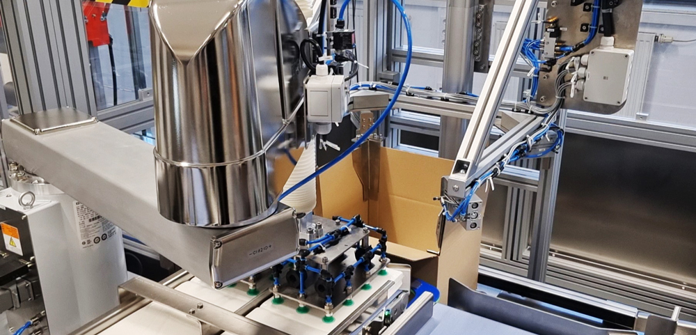 Automatisierte-verpackungsanlage-fuer-maximale-flexibilitaet-item-blog-bild-im-beitrag-3 Un robot scara con pinzas transfiriendo un paquete de papel a una caja